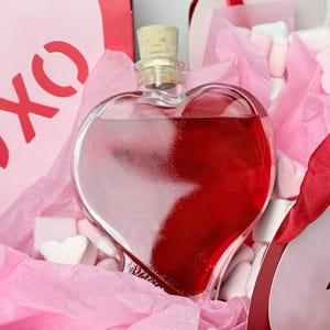 May include: A heart-shaped glass bottle filled with red liquid, sealed with a cork stopper. The bottle is nestled in pink tissue paper and heart-shaped marshmallows. A red gift bag with "XO" is in the background.