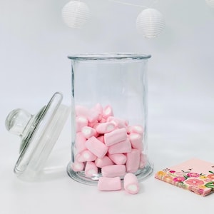 May include: A clear glass jar filled with pink marshmallows, lid off to the side. The jar is on a white surface, with a floral napkin and white paper lanterns in the background. The marshmallows are pink.