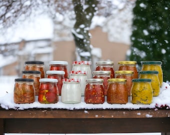 May include: A collection of small glass jars filled with various red and yellow pickled vegetables. The jars are arranged in two rows on a wooden surface covered in snow.