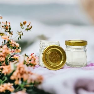 May include: Two clear glass jars with gold lids. The jars are sitting on a white surface with pink flowers in the background.
