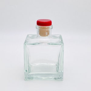 Op de afbeelding: Een heldere glazen fles met een vierkante vorm en een rode kurk. De fles is leeg en klaar om gevuld te worden.