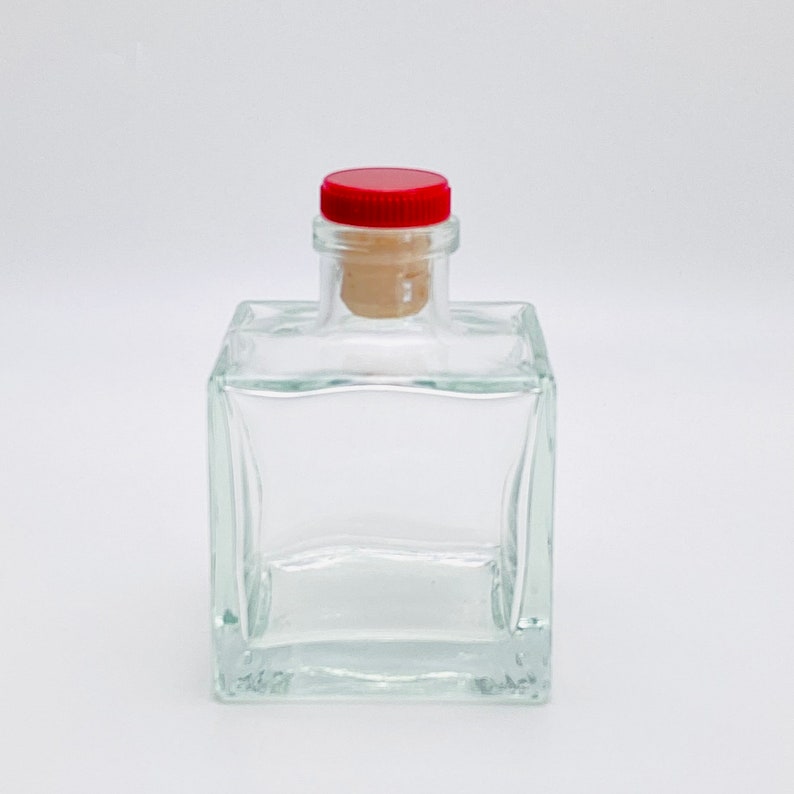 Op de afbeelding: Een heldere glazen fles met een vierkante vorm en een rode kurk. De fles is leeg en klaar om gevuld te worden.