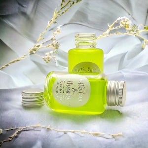 Op de afbeelding: Twee kleine glazen flesjes gevuld met een heldergroene vloeistof. De flesjes hebben witte etiketten met gouden letters die "with love & thanks" en "little love" zeggen.