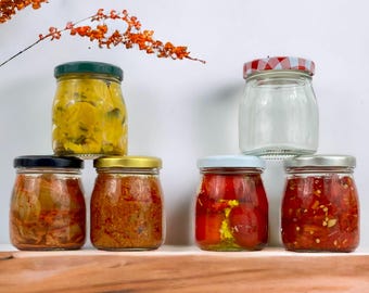 May include: A collection of glass jars filled with various preserved foods. The jars have different colored lids, including green, black, gold, silver, and a red and white checkered pattern. The contents appear to be pickled vegetables and sauces.