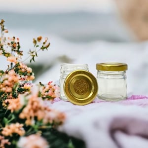 May include: Two small glass jars with gold lids. The jars are empty and sitting on a white fabric surface.