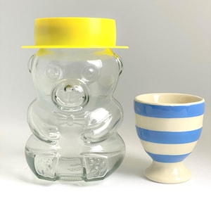 May include: A clear glass bear-shaped jar with a yellow lid and a small, striped blue and white egg cup. The jar has a bow tie detail. The egg cup has a pedestal base.