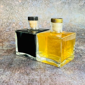 Op de afbeelding: Twee vierkante glazen flessen met kurkstoppen. De fles links is gevuld met een donkere vloeistof en de fles rechts is gevuld met een lichtgele vloeistof.