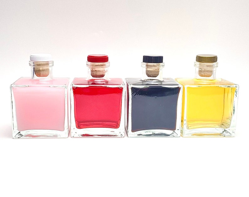 Op de afbeelding: Vier vierkante glazen flessen met kurkstoppen en verschillende gekleurde vloeistoffen. De flessen zijn in een rij van links naar rechts gerangschikt: roze, rood, zwart en geel.