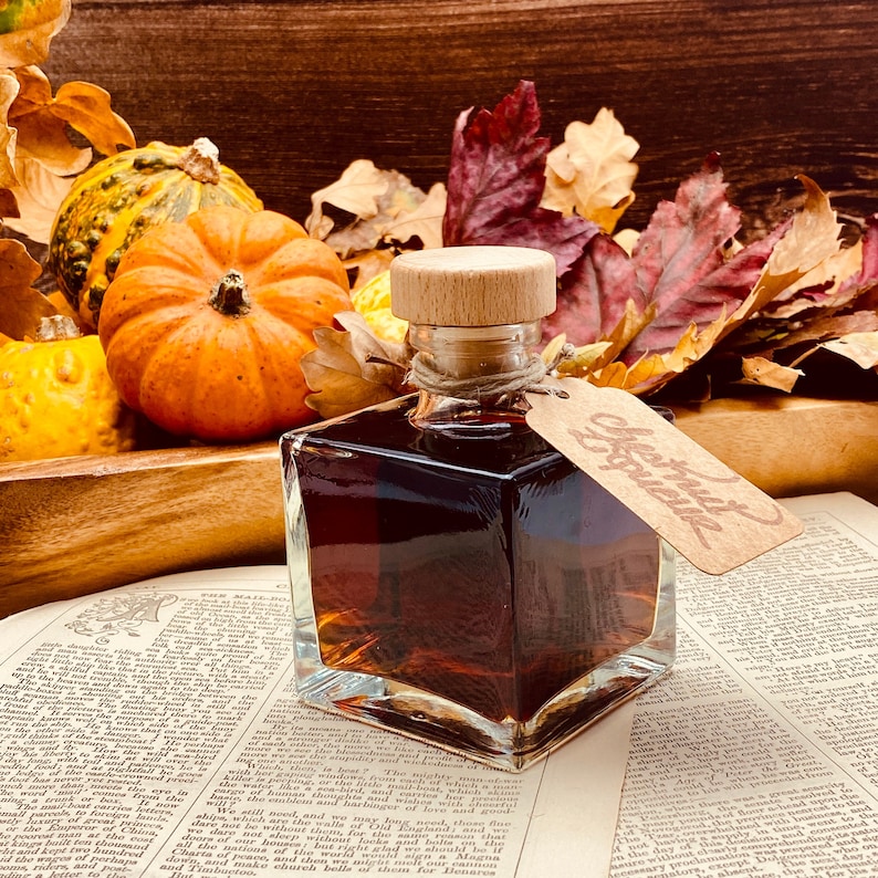 Op de afbeelding: Een vierkante glazen fles met een houten stop en een bruine vloeistof erin. De fles staat op een boek met een bruine leren kaft. De fles heeft een bruin papieren label eraan bevestigd met de tekst "Cherry & Spice". De fles is omringd door herfstbladeren en kleine pompoenen.