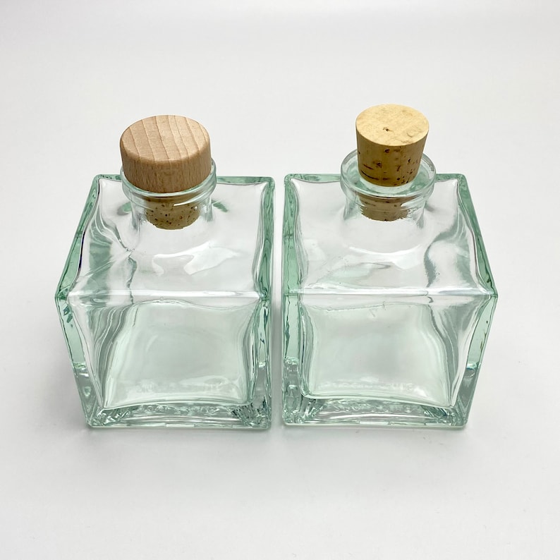 Op de afbeelding: Twee vierkante glazen flessen van helder glas met kurken stoppen. De flessen zijn leeg en hebben een getextureerd oppervlak.