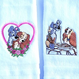 May include: Two white kitchen towels with embroidered images of Lady and the Tramp. One towel shows Lady and Tramp cuddling inside a pink heart with roses. The other towel shows Lady and Tramp sharing a plate of spaghetti.