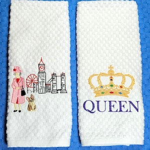 May include: Two white kitchen towels with embroidered designs. The first towel features a cartoon of the Queen of England, Big Ben, the London Eye, and Tower Bridge. The second towel features a gold crown and the word "QUEEN" in purple.