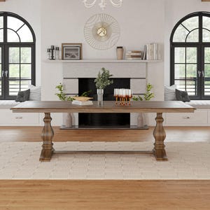 May include: A large, rectangular, wooden dining table with a trestle base. The table is in a light brown wood finish and has a distressed look. The table is set with a white rug underneath and is in a room with large windows and a fireplace.
