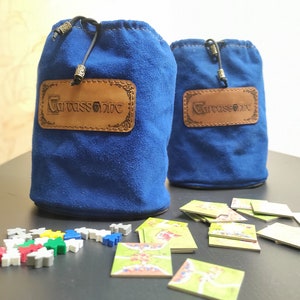 Op de afbeelding: Twee blauwe fluwelen zakken met bruine leren pleisters waarop "Carcassonne" staat. De zakken liggen op een zwart oppervlak met een stapel kleine, kleurrijke spelstukken en een stapel speltegels.