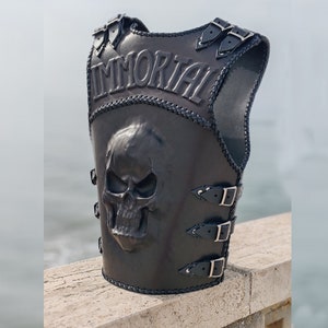 May include: Black leather vest featuring a skull design and the word "IMMORTAL" embossed on the front. The vest has adjustable straps and buckles.