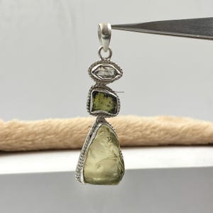 Może przedstawiać: Srebrny wisiorek z trzema zielonymi i przezroczystymi kamieniami szlachetnymi. Wisiorek jest ozdobiony sznurkowym obramowaniem wokół każdego kamienia.