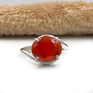 Puede incluir: Un anillo de plata con una gran piedra redonda de cornalina roja. El anillo tiene un diseño simple y elegante.
