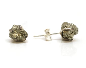 Op de afbeelding: Een paar oorknopjes met ruwe, metaalachtig goudkleurige pyriet mineraalclusters. Elke steen met een ruwe textuur is stevig bevestigd op een zilverkleurige metalen stift met een bijpassende vlindersluiting, wat een natuurlijke, onregelmatige vorm laat zien.
