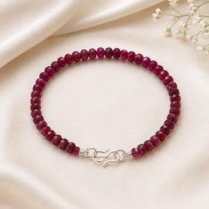 May include: A beaded bracelet featuring small, faceted, deep magenta-red gemstone beads arranged in a circular pattern. The bracelet is secured with a silver-coloured metal S-hook clasp, which connects the two ends of the beaded strand together.