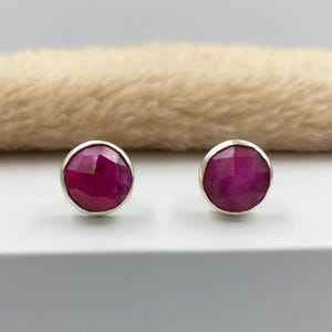 Peut inclure: Une paire de boucles d'oreilles rondes. Chaque boucle d'oreille présente une pierre précieuse rose foncé à facettes sertie dans une lunette de couleur argentée. Les boucles d'oreilles sont présentées sur une surface blanche avec un arrière-plan flou et beige.