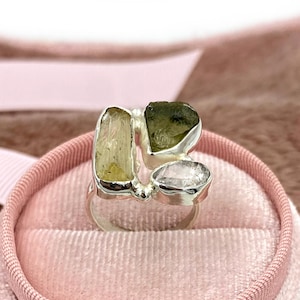 Puede incluir: Un anillo de plata con tres piedras preciosas en bruto. El anillo presenta una piedra verde claro, una piedra verde oscuro y una piedra de cuarzo transparente.