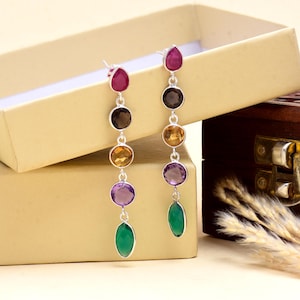 Könnte beinhalten: Ein Paar lange, baumelnde Ohrringe mit silbernem Finish. Jeder Ohrring hat eine Reihe farbiger Edelsteine, darunter Rubin, Braun, Gelb, Amethyst und Grün, in Tropfen- und Kreisform. Die Ohrringe werden in einer offenen, beigefarbenen Schmuckschatulle präsentiert.