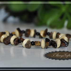 May include: A brown, black, and white beaded bracelet with a tiger's eye stone design. The bracelet is on a white surface with a green plant in the background. The text "T.Berrys.Gifts" is visible in the lower right corner of the image.