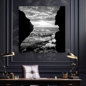 May include: A black and white photograph of a seascape viewed through a rock formation. The photo is hanging on a wall in a dark office with a leather office chair and a desk with two lamps.