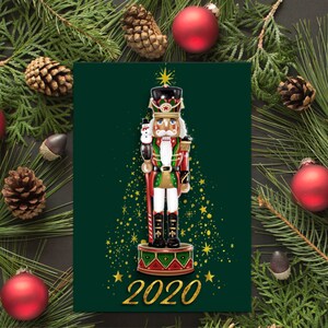 May include: A green Christmas card with a nutcracker soldier standing in front of a gold glitter Christmas tree. The nutcracker is wearing a red and green uniform and holding a candy cane. The card has the year 2020 written in gold glitter.