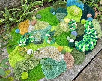 以下が含まれることがあります： 緑、茶色、青のふわふわした糸でできた、さまざまな質感のラグ。ラグは丘と谷のある風景の形をしています。