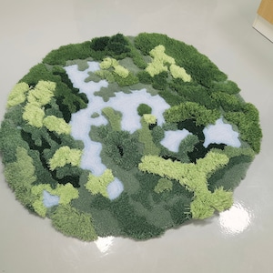 May include: A round, green and blue rug with a textured, fuzzy surface. The rug is designed to look like a landscape with green hills and blue rivers.