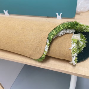 May include: A rolled-up rug with a green and white fringe. The rug has a beige backing and is sitting on a light wood surface.