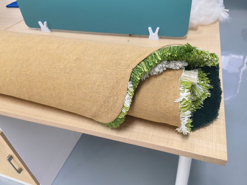 May include: A rolled-up rug with a green and white fringe. The rug has a beige backing and is sitting on a light wood surface.