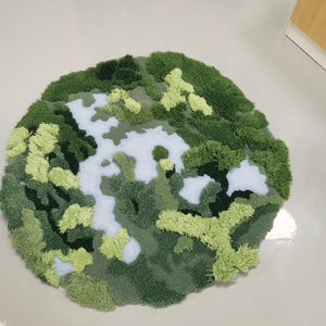 May include: A round, green and white rug with a textured, fuzzy surface. The rug is designed to look like a landscape with green hills and a white river.