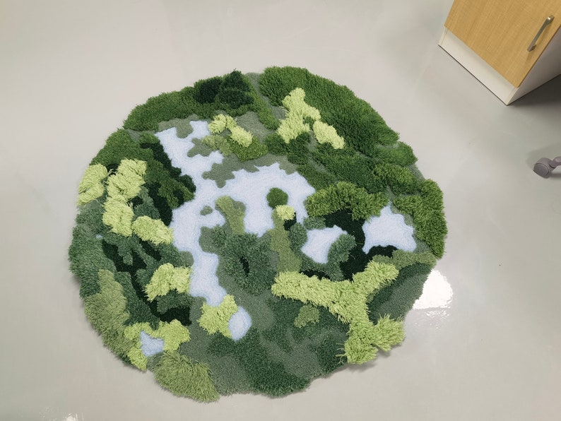 May include: A round, green and white rug with a textured, fuzzy surface. The rug is designed to look like a landscape with green hills and a white river.