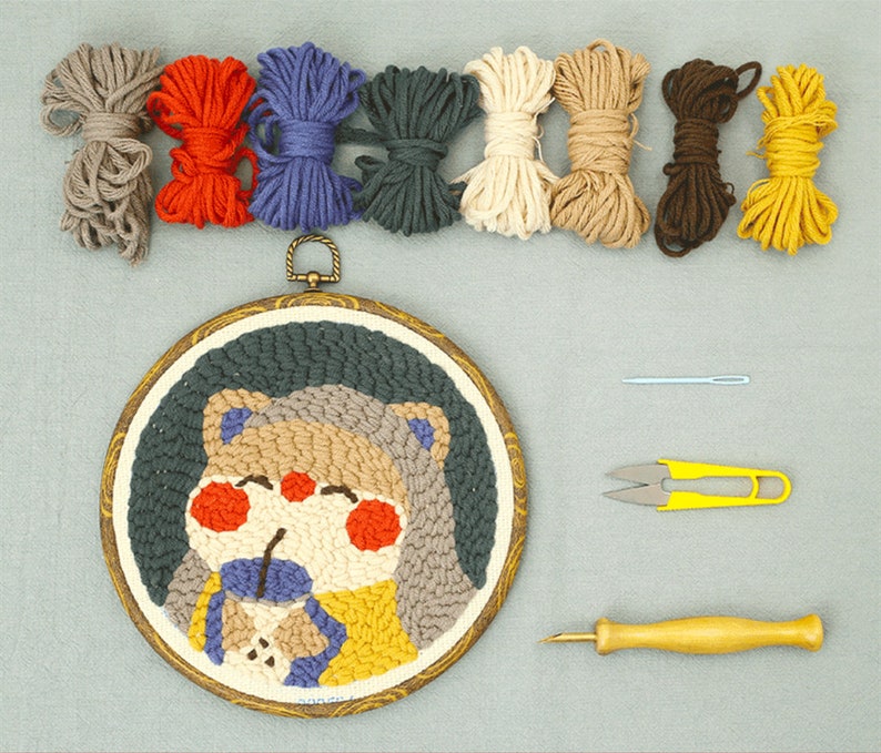 May include: A craft kit with a round, framed textile art piece depicting a cartoon bear, alongside bundles of coloured yarn, a needle, scissors, and a punch needle tool. The bear art features a blue, yellow, and brown colour scheme.