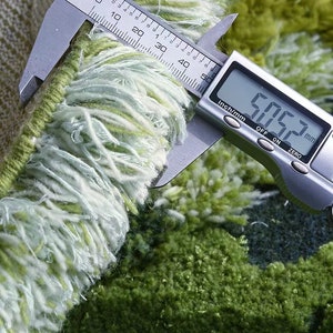 May include: Close-up of a green and white rug fringe being measured with a digital caliper. The caliper reads 5.063 mm.