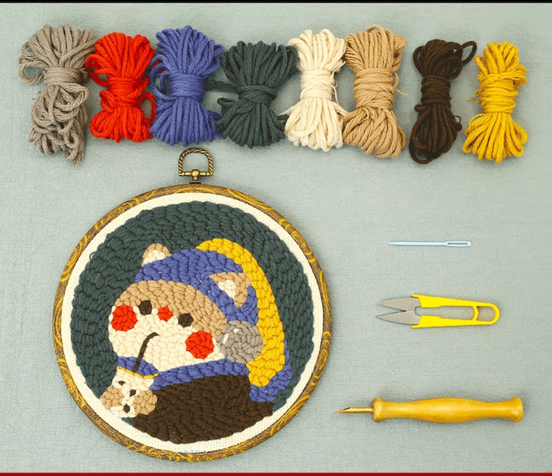 May include: A craft kit featuring a round, framed punch needle art piece with a cartoon character design, alongside bundles of colourful yarn, a needle, scissors, and a punch needle tool. Yarn colours include grey, red, blue, green, cream, brown, and yellow.