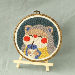 May include: A circular embroidered artwork depicting a cartoon bear. The bear has a light brown face, red cheeks, and wears blue and yellow clothing. The embroidery is set in a wooden hoop with a patterned border, displayed on a small wooden easel.