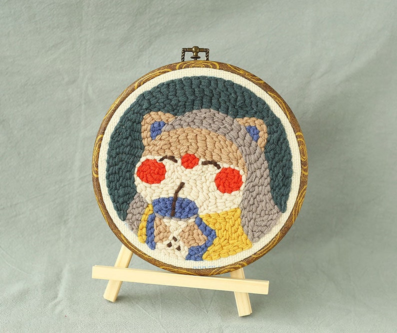 May include: A circular embroidered artwork depicting a cartoon bear. The bear has a light brown face, red cheeks, and wears blue and yellow clothing. The embroidery is set in a wooden hoop with a patterned border, displayed on a small wooden easel.