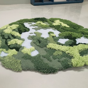 May include: A round, green and white rug with a textured, fluffy surface. The rug is designed to look like a landscape with green hills and white streams.