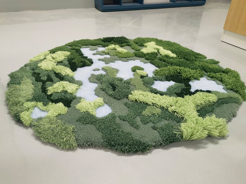 May include: A round, green and white rug with a textured, fluffy surface. The rug is designed to look like a landscape with green hills and white streams.