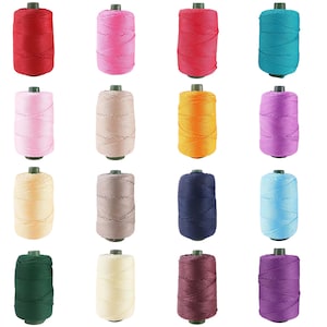May include: A collection of sixteen cylindrical spools of thread arranged in a four-by-four grid. Each spool features a distinct solid color, including red, pink, magenta, teal, light pink, beige, orange, purple, cream, gray, navy, blue, green, off-white, maroon, and violet.