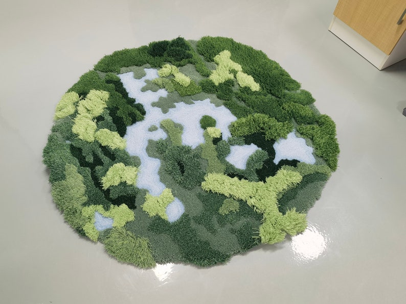 May include: A round, green and blue rug with a textured, fuzzy surface. The rug is designed to look like a landscape with green hills and blue rivers.