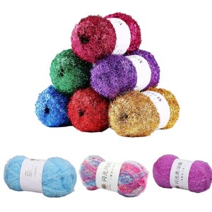 Può includere: Una collezione di gomitoli di lana colorati in varie tonalità, tra cui rosso, verde, blu, viola e oro. Alcuni gomitoli sono impilati, altri sono esposti singolarmente. Ogni gomitolo ha un'etichetta bianca. La lana appare morbida e soffice.
