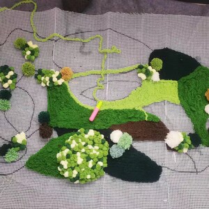 May include: A green, brown, and white rug made of pom-poms on a white mesh canvas. The rug is shaped like a stylized island with a winding path.