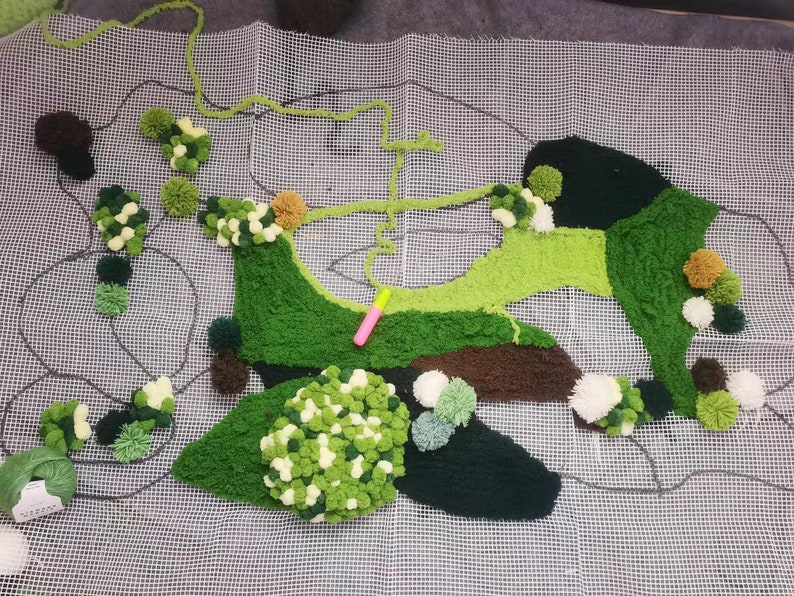 May include: A green, brown, and white rug made of pom-poms on a white mesh canvas. The rug is shaped like a stylized island with a winding path.