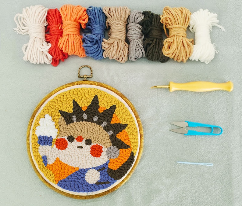 May include: A punch needle kit with a cartoon character design. The kit includes a wooden embroidery hoop, colourful yarn skeins, a punch needle tool, scissors, and a needle. The finished piece has a yellow background and a cartoon character with a crown.