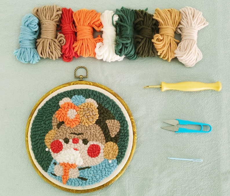 May include: A punch needle embroidery kit with a cartoon character design. The kit includes a round embroidery hoop, colourful yarn skeins, a punch needle tool, scissors, and a needle. The design features a character with a flower.