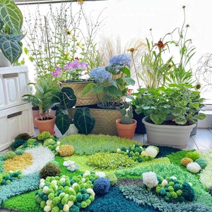 May include: A green, blue, and white rug with a textured, fluffy surface. The rug is decorated with pom-poms and resembles a garden scene. Several potted plants are visible in the background.